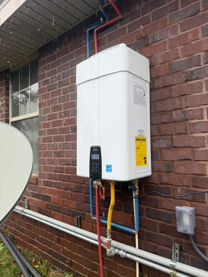 Outdoor tankless water heater mounted on a brick wall with red and blue water lines and a yellow gas connector