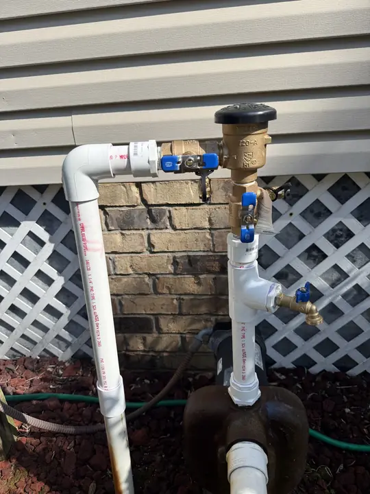 New brass backflow preventer with blue-handled shutoff valves on white PVC pipe beside a house