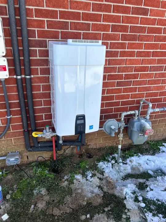 Outdoor tankless water heater on a brick wall with insulated pipes and gas meter, snow on the ground